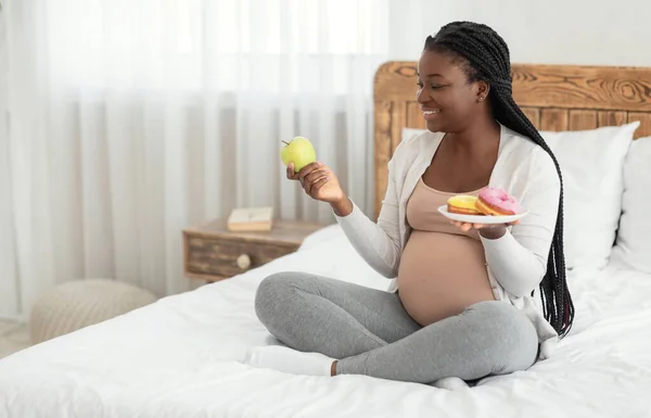 Nutrición prenatal. Mujer embarazada negra eligiendo entre manzana y rosquillas en casa — Foto de Stock