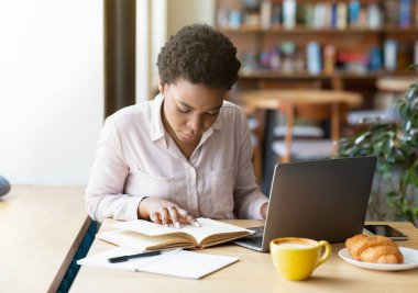 Kafede laptopun önünde kitap okuyan zeki siyah kadın sınava hazırlanıyor ya da iş projesi üzerinde çalışıyor.