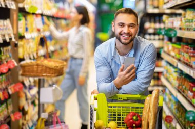 Cep telefonlu genç adam hipermarkette alışveriş yapıyor.