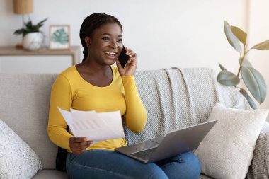 Siyah serbest çalışan kadın cep telefonuyla konuşuyor ve evdeki kağıtlarla çalışıyor.