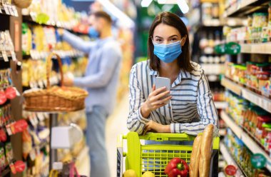 Hipermarkette akıllı telefon alışverişi yapan maskeli bir kadın.