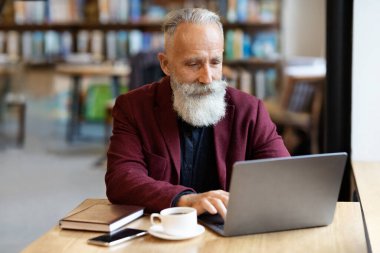 Kafede dizüstü bilgisayar kullanan, kahve içen gri saçlı iş adamı.