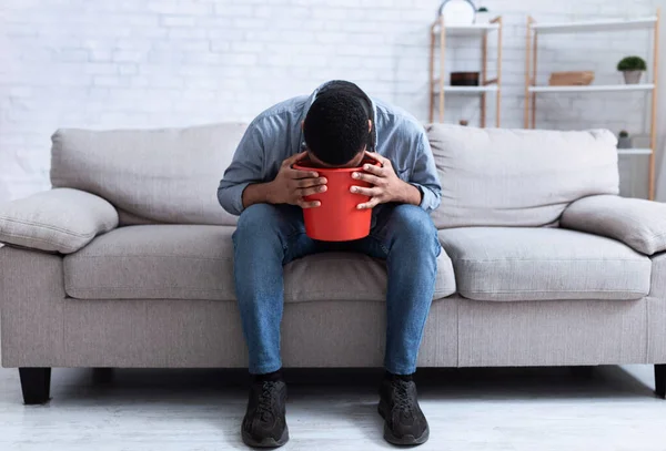 Hombre africano vomitando en un cubo sentado en un sofá en casa — Foto de Stock