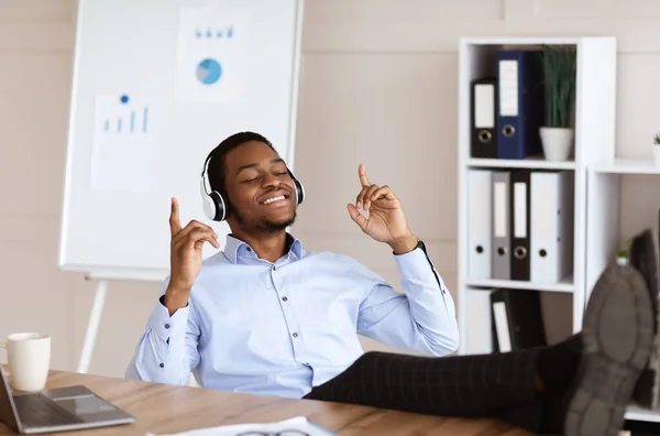 Joven hombre de negocios negro escuchando música con piernas en la mesa —  Fotos de Stock