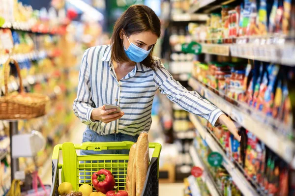 Cep telefonu tutan maskeli kadın dükkânda durup ürünler alıyor.