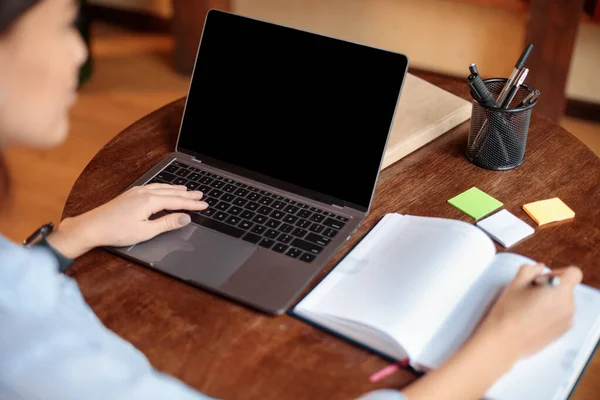 Kvinne som sitter ved skrivebordet med en bærbar pc med tom skjerm, som skriver – stockfoto