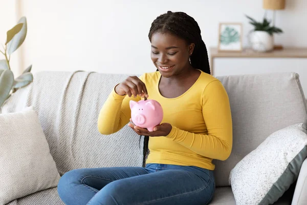 Mulher negra colocando moeda no banco porquinho, sentado no sofá em casa — Fotografia de Stock