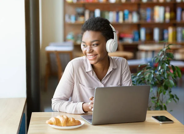 Atractiva mujer afroamericana con auriculares que utilizan ordenador portátil para la comunicación en línea en la cafetería — Foto de Stock