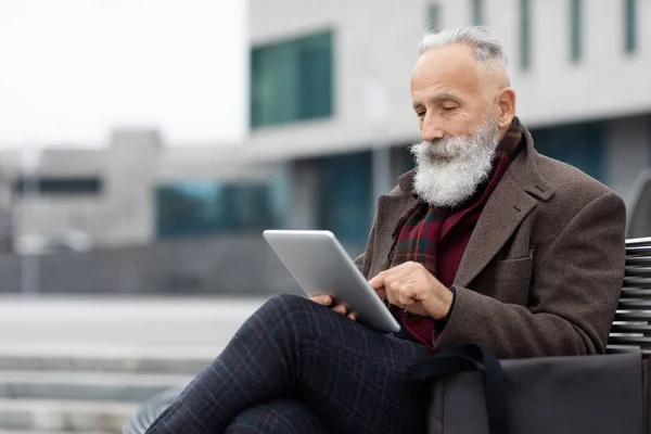 Tarz sahibi gri saçlı yaşlı adam bankta oturuyor, dijital tablet kullanıyor. — Stok fotoğraf