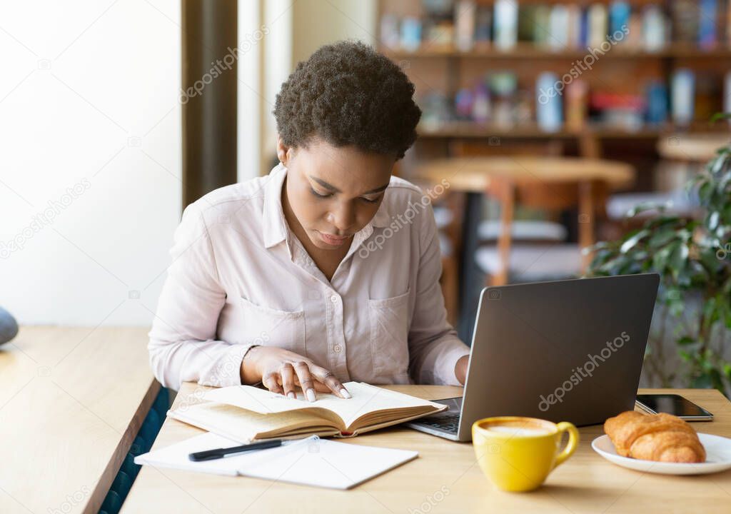 Smart black woman reading book in front of laptop at cafe, getting ready for exam or working on business project
