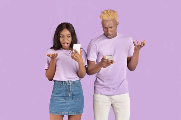 Joven mujer negra su novio mirando en sus teléfonos inteligentes en shock en violeta fondo del estudio — Foto de Stock