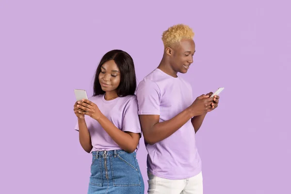 Jovem senhora negra com seu namorado usando telefones celulares no fundo do estúdio lilás — Fotografia de Stock