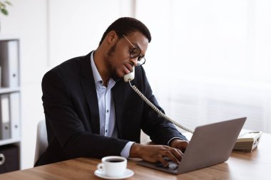 Meşgul siyah yönetici telefonla konuşuyor ve dizüstü bilgisayar kullanıyor.