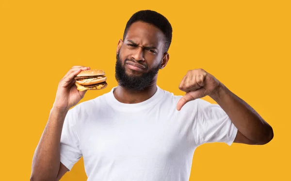 Preto homem com hambúrguer gestando polegares para baixo de pé no fundo amarelo — Fotografia de Stock
