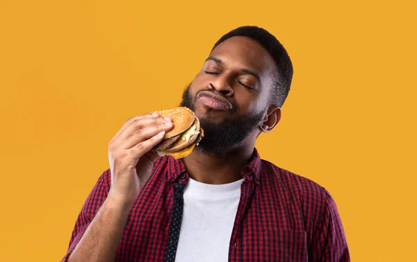 Africano Millennial Guy Smelling Burger em pé no fundo do estúdio amarelo — Fotografia de Stock