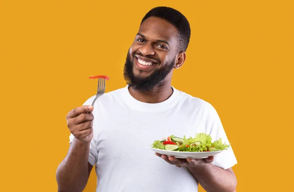 Alegre afro-americano cara comer salada posando sobre fundo amarelo — Fotografia de Stock