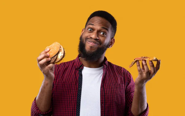 Chico negro feliz sosteniendo hamburguesa y pizza sobre fondo amarillo —  Fotos de Stock