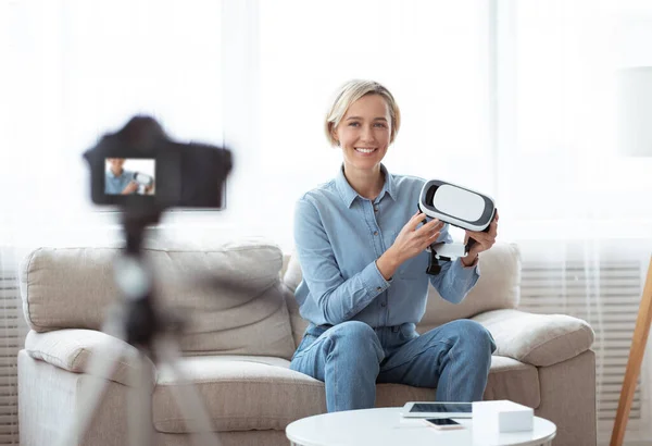 Young female vlogger holding VR headset, showing innovative device, recording video for her technology blog on camera