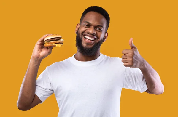Africano Guy Segurando Burger Gesturing Thumbs-Up em fundo de estúdio amarelo — Fotografia de Stock