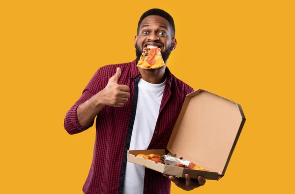 Preto cara com pizza no boca gesturing polegares para cima, fundo amarelo — Fotografia de Stock