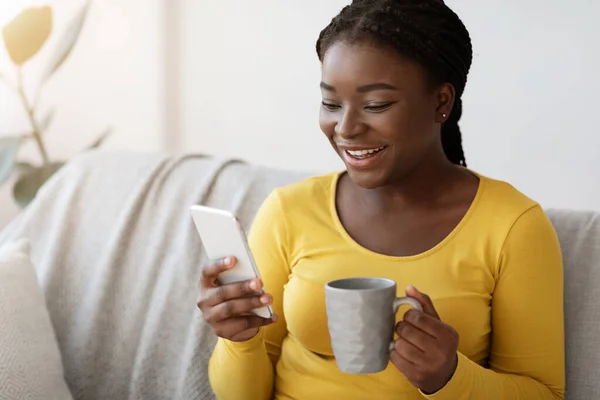 Ευχάριστο μήνυμα. Ευτυχισμένη μαύρη γυναίκα χρησιμοποιώντας Smartphone και πίνοντας καφέ στο σπίτι — Φωτογραφία Αρχείου