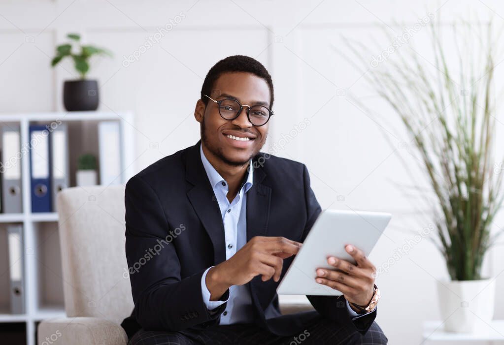Smiling black manager in suit holding digital tablet, office interior