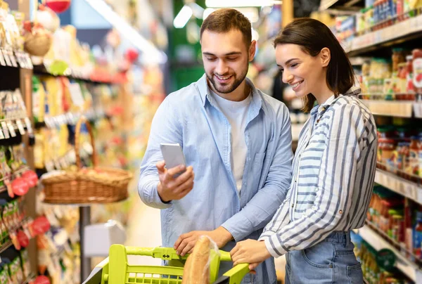 슈퍼마켓에서 카트와 스마트폰 쇼핑을 하고 있는 젊은 부부 — 스톡 사진