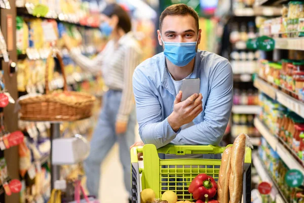 Guy in face mask με smartphone ψώνια στο hypermarket — Φωτογραφία Αρχείου