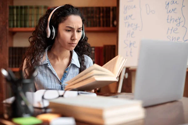 Mujer sentada en el escritorio, tocando usando auriculares, usando computadora, leyendo libro —  Fotos de Stock