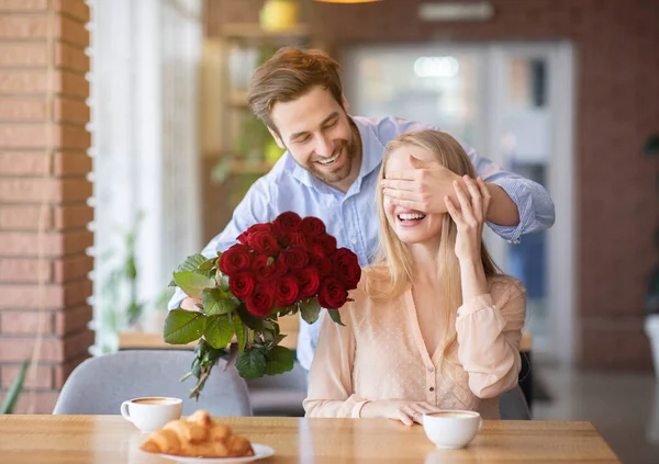 Surpresa romântica. Jovem carinhoso dando buquê de flores para sua amada mulher, cobrindo seus olhos no café da cidade — Fotografia de Stock