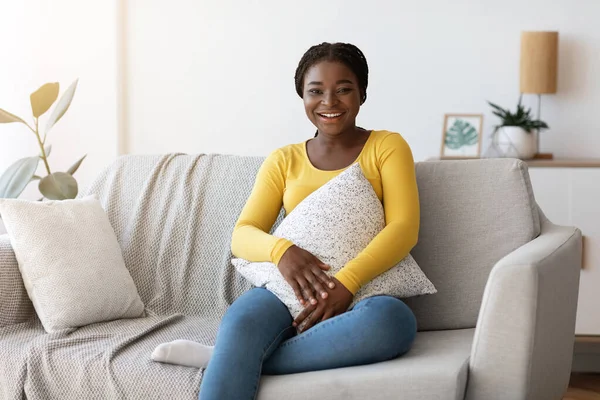 Dia em casa. Sorrindo mulher negra sentada no sofá confortável com travesseiro — Fotografia de Stock