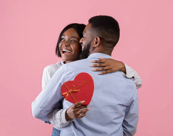 Mulher negra excitada segurando caixa de presente em forma de coração e abraçando seu namorado no fundo do estúdio rosa — Fotografia de Stock