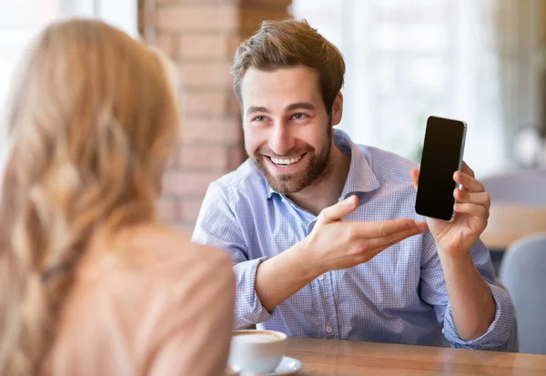 Positivo ragazzo millenario che mostra il suo cellulare fidanzata con schermo bianco alla caffetteria, mockup per il design — Foto Stock