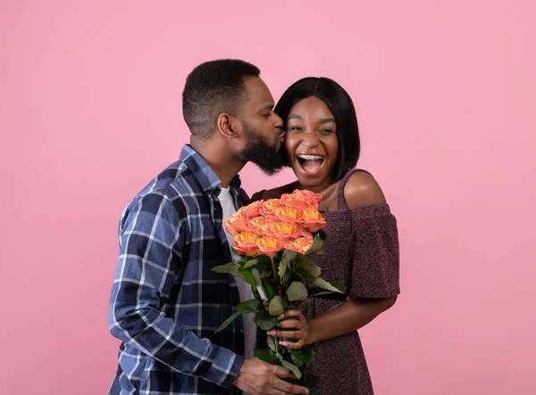 Любящий афроамериканец целует свою счастливую девушку и дарит ей букет роз на фоне розовой студии — стоковое фото