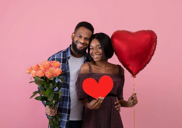 Dia dos Namorados. Jovem casal preto com coração vermelho, balão e buquê de rosas posando no fundo do estúdio rosa — Fotografia de Stock