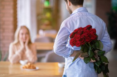 Arkasında bir buket kırmızı gülle romantik genç adam kafedeki kız arkadaşına sürpriz yapıyor.