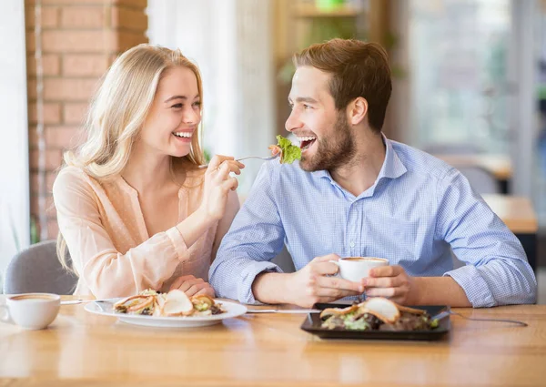 Lovely young woman feeding her boyfriend with fresh salad at coffee shop