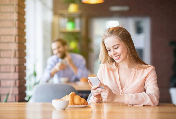Vonzó fiatal hölgy böngészés web okostelefon a kávézóban, fiatal férfi megfigyeli őt hátulról, másolja tér — Stock Fotó