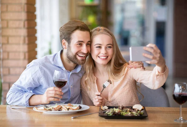 Pozytywna młoda para jedząca razem lunch w restauracji, mówiąca selfie podczas posiłku — Zdjęcie stockowe