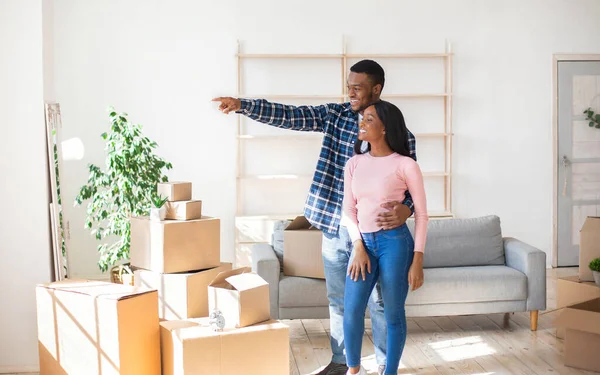 Africano americano cara mostrando algo para sua namorada feliz, de pé entre as caixas em sua nova casa, espaço livre — Fotografia de Stock