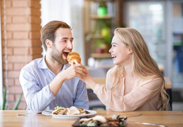 Loving young woman feeding yummy croissant to her boyfriend at urban cafe