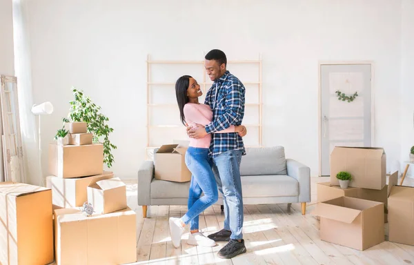 Mooie zwarte vrouw en haar vriend kijken elkaar aan en knuffelen in kamer gevuld met kartonnen dozen op verhuisdag — Stockfoto