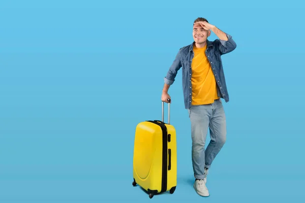 Pełna długość portret szczęśliwy mężczyzna podróżnik z bagażem patrząc na pustą przestrzeń nad niebieskim tle studio — Zdjęcie stockowe