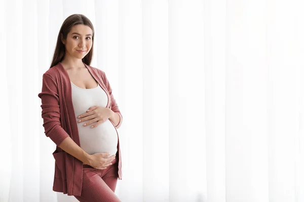 Retrato da mulher grávida bonita que está perto da janela em casa, abraçando a barriga — Fotografia de Stock