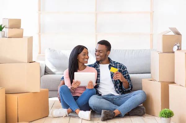 Preto casal de compras de bens domésticos on-line no dia em movimento, sentado com laptop e cartão de crédito entre caixas de papelão — Fotografia de Stock