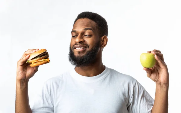 Homem africano escolhendo entre hambúrguer e maçã de pé no estúdio — Fotografia de Stock