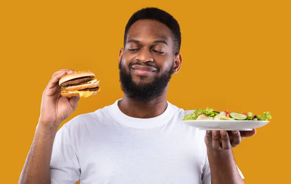 Homem com fome escolhendo entre hambúrguer e salada em fundo amarelo — Fotografia de Stock