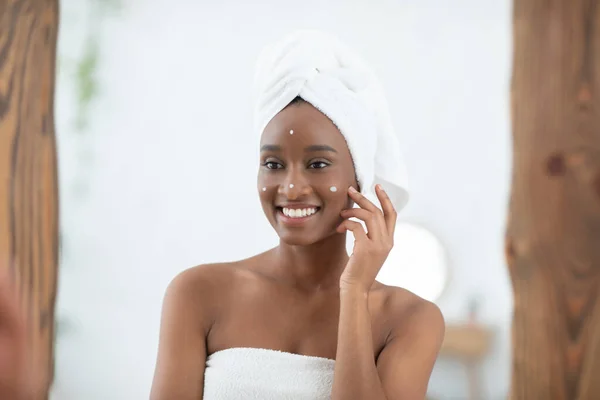 Souriante jeune femme afro-américaine avec une serviette blanche sur la tête enduit son visage de crème — Photo