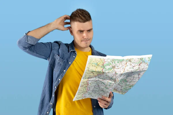 Homem frustrado examinando mapa e coçando a cabeça, sentindo-se perdido, tendo problemas para encontrar seu destino, fundo azul — Fotografia de Stock