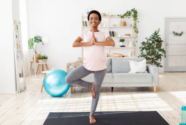 Bekleyen genç bayan sabah yoga pratiği yapıyor, ağaç duruşu yapıyor, evde kendini huzurlu hissediyor.